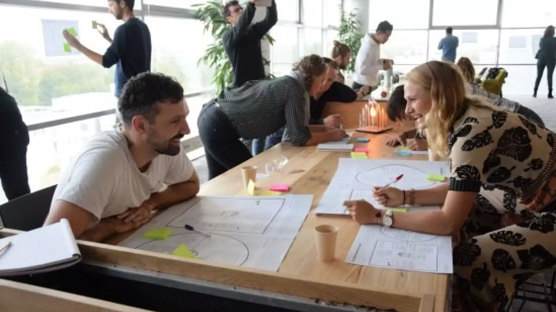 Mensen staan en zitten aan een lange tafel en brainstormen samen. Op de tafel liggen witte vellen met hierop stickers en schrijfwerk.