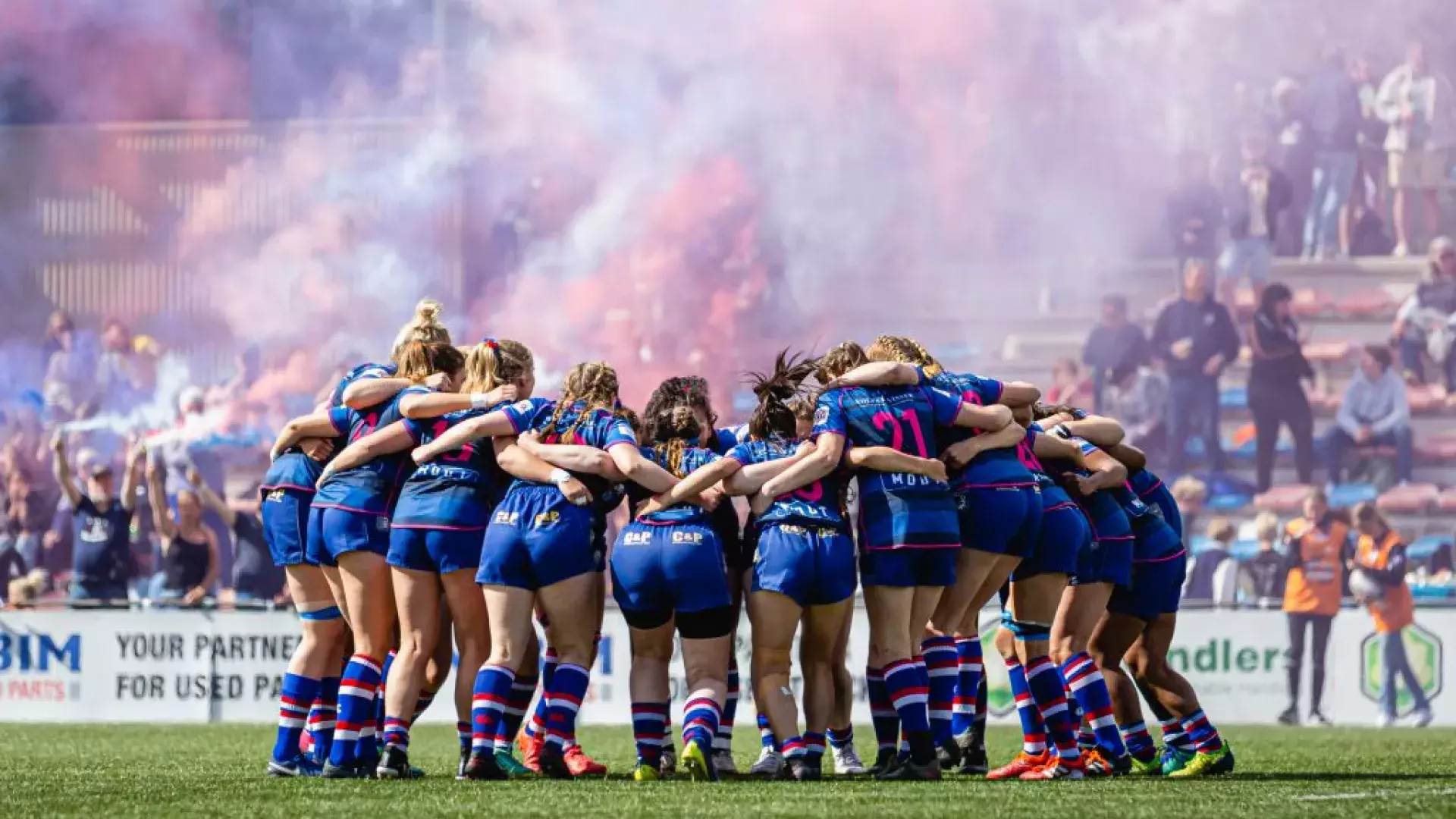 groep vrouwelijke rugby'ers die midden op het veld de winst vieren. Ze staan in een kring en houden elkaar vast. Op de achtergrond zie je een volle tribune met fakkels en rook