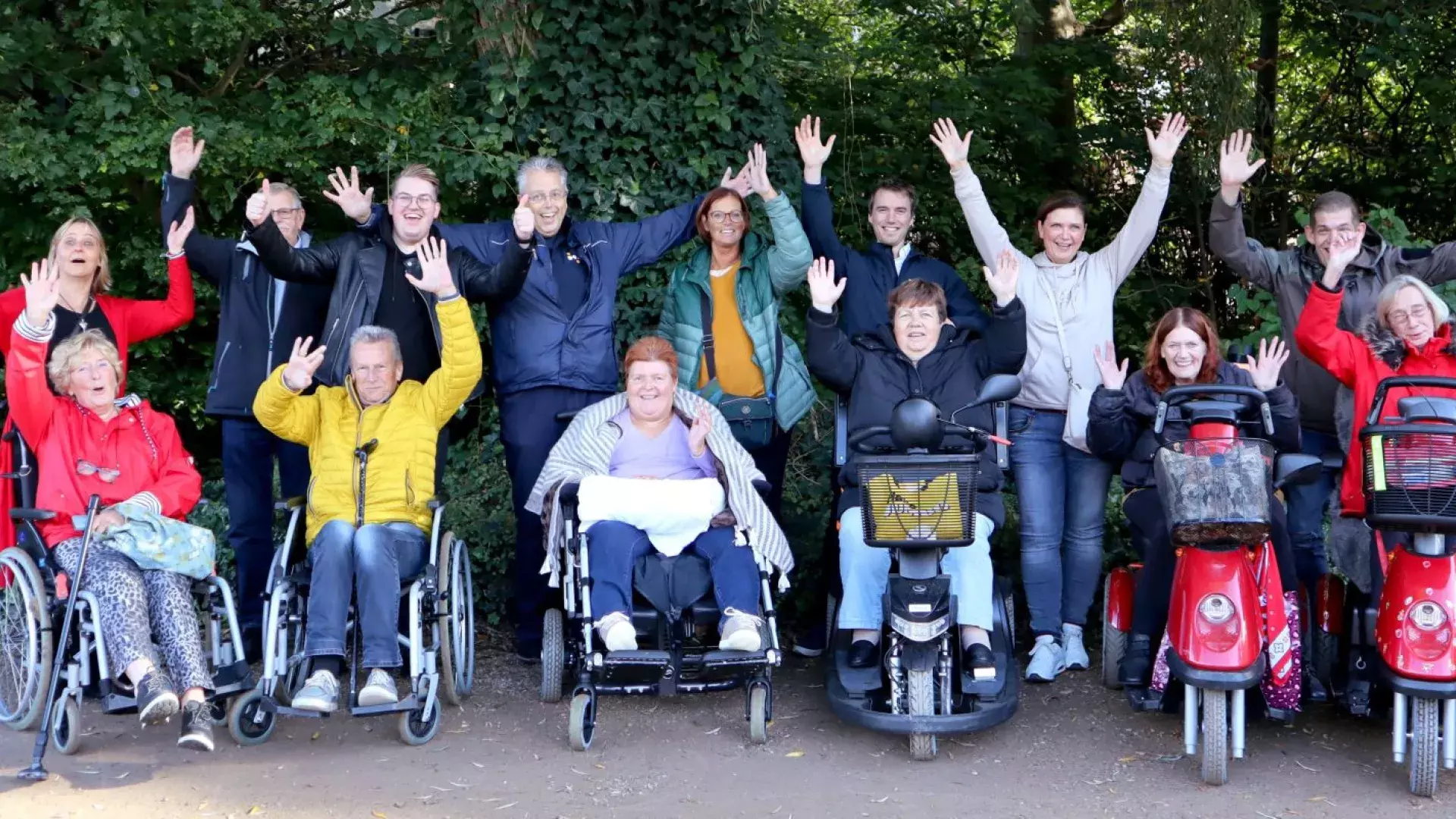 teamuitje van een organisatie. Op de voorgrond zijn mensen in een rolstoel, daarachter staan medewerkers van een organisatie. Allemaal lachen ze naar de camera en hebben hun handen omhoog.
